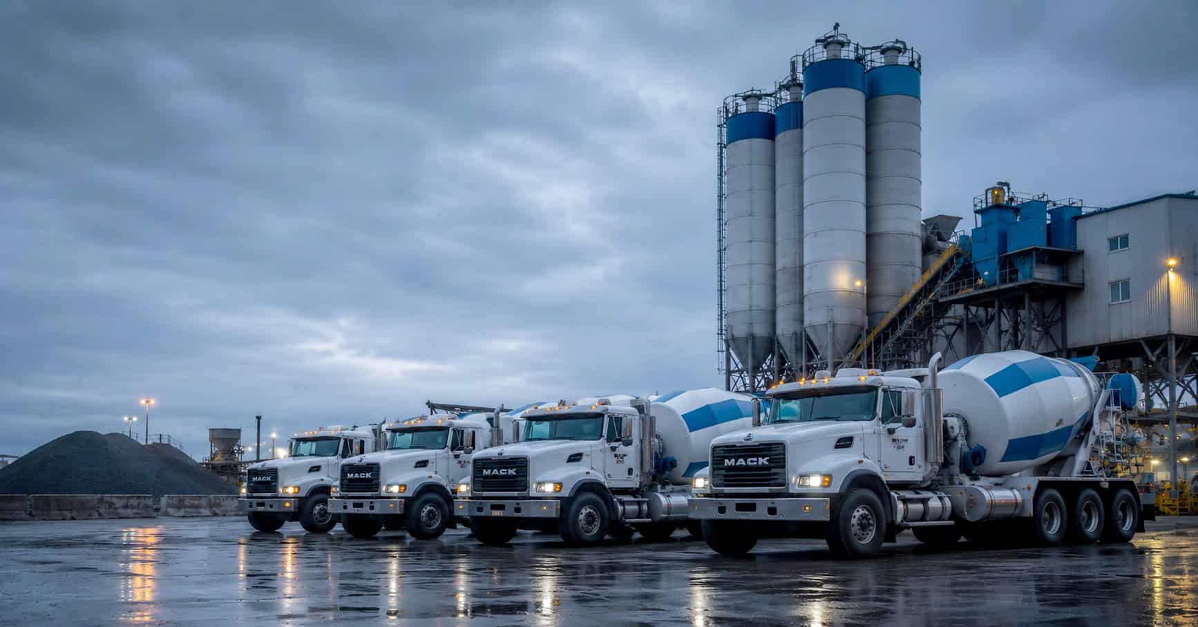 Ready-mix concrete trucks lined up at a plant with silos at dusk
